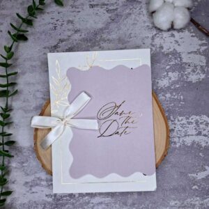 کارت دعوت عروسی یاس رنگ یاسی با پاکت طلاکوب Save The Date در دست عروس در فضای باغ
